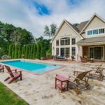 Poolside view of a Salt Box style barn home with grand windows beautifully matching, smaller salt box poolhouse by Harvest Moon