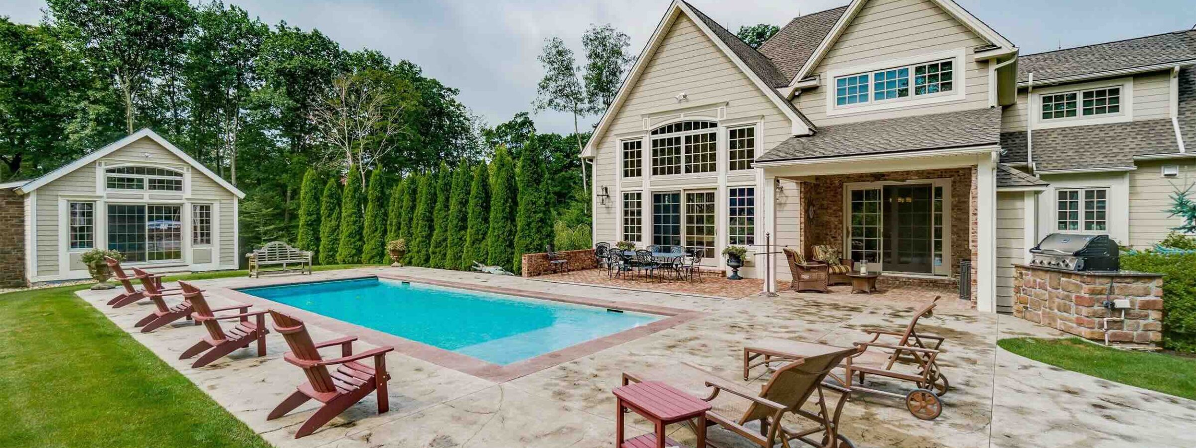 Poolside view of a Salt Box style barn home with grand windows beautifully matching, smaller salt box poolhouse by Harvest Moon